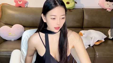 Media: Video of an East Asian woman with long black hair, fair skin, and a slender physique, wearing a black sleeveless top, sitting on a brown leather couch. Background includes stuffed animals, including a pink bear and a yellow duck.