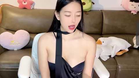 Media: A video of an Asian woman with long black hair, wearing a black strapless dress, sitting on a brown leather couch with stuffed toys. She has a sensual expression, leaning back with one hand on her chest.