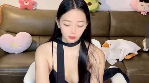 Media: A video of an East Asian woman with long black hair, wearing a black choker and revealing black dress, sitting on a green couch with plush toys.