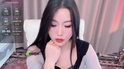 Media: Video of a young Asian woman with long black hair, wearing a sheer black top, sitting with a contemplative expression. Background includes a white chair and a blurred, beige curtain.