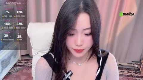 Media: Video of an East Asian woman with long black hair, light skin, wearing a black and white plaid shirt, sitting on a white couch, with a live streaming overlay displaying viewer count.