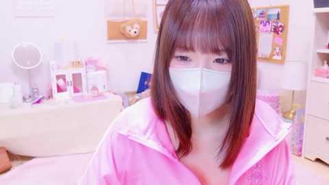 Media: Video of an East Asian woman with long, straight brown hair, wearing a pink robe and white face mask, sitting in a brightly lit, cluttered bedroom with toys, a mirror, and a stuffed bear.