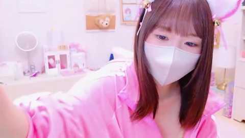 Media: Video of an Asian woman with straight brown hair, wearing a pink satin robe, white mask, and pink bunny ears, taking a selfie in a brightly lit, pink-themed bedroom with white furniture.