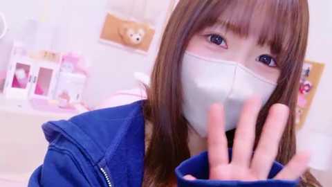 Media: A video of an Asian woman with long brown hair, wearing a blue jacket and white face mask, waving in a soft-focus, pastel-toned room with plush toys and a teddy bear.