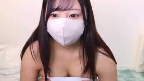 Media: Video of an East Asian woman with long black hair, wearing a white face mask and a strapless top, seated on a bed with white sheets, in a minimalistic, soft-colored room.