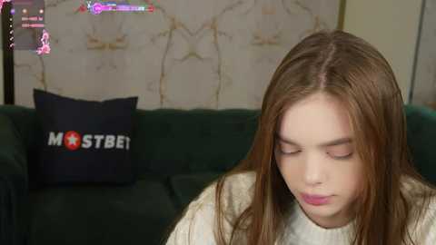 Media: Video of a young woman with long brown hair, wearing a beige sweater, sitting on a green couch with a dark blue pillow labeled \"MOSTBET.\" The background shows a floral wallpaper and a gaming setup.