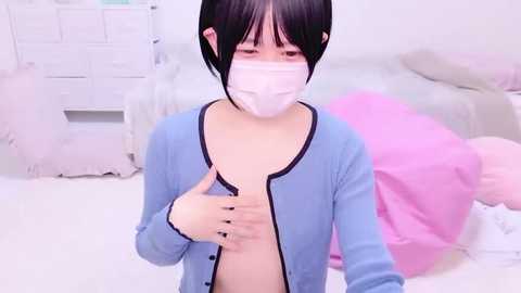 Media: A video of an East Asian woman with short black hair, wearing a white mask, blue cardigan, and a white shirt, standing in a white room with pink bedding and a white dresser.