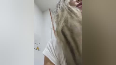 Media: A video captures a person with long, wavy blonde hair, wearing a white T-shirt, partially obscured by a vertical object, likely a wall or door, in a white-walled room.