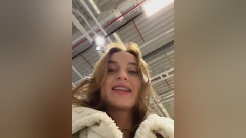 Media: Video of a smiling young woman with light brown hair and fair skin, wearing a white fuzzy jacket, inside a dimly lit industrial warehouse with exposed pipes and beams.