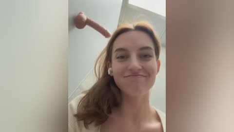 Media: Video of a smiling young woman with medium-length brown hair, wearing a beige sweater, standing in a bathroom with a mounted dildo on the wall.