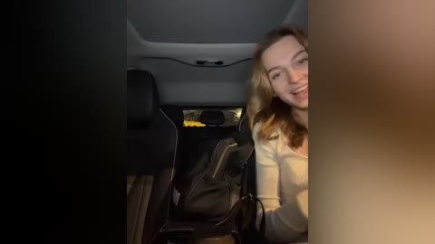 Media: Video of a smiling young woman with blonde hair and fair skin, wearing a light-colored top, sitting in the backseat of a dark car, with a black backpack on the seat beside her.