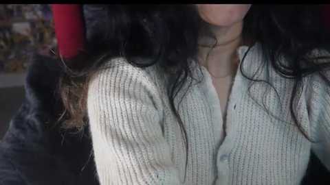 Media: A video shows a woman with long, wavy black hair, wearing a light grey knit sweater with a V-neck, and a delicate necklace. The background is blurred, featuring a red object and a hint of a room with colorful patterns.