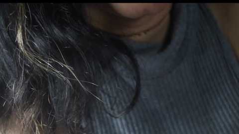 Media: A close-up video of a person's head, showing a section of their dark hair with light streaks and a gray ribbed sweater. The background is out of focus.