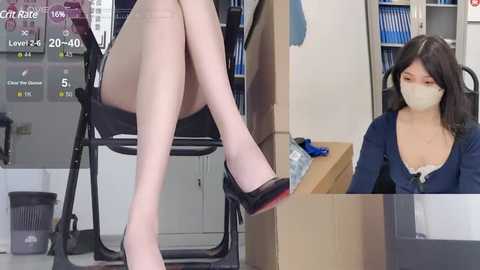 Media: Video of a woman wearing a face mask, seated at a desk, with a pair of legs in black high heels in the foreground, in a modern office setting.