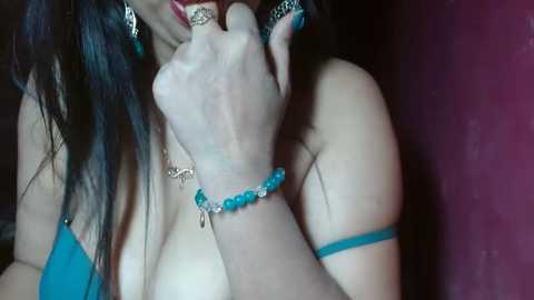 Media: A close-up video of a woman with long black hair, fair skin, and a turquoise bracelet, biting her finger, wearing a blue top.