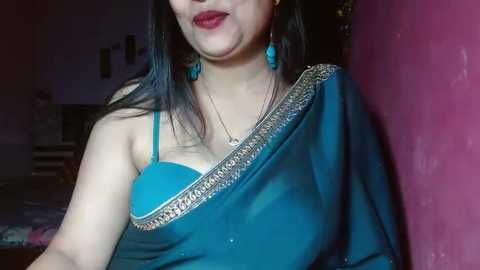 Media: A video of a South Asian woman with fair skin, long black hair, and red lipstick, wearing a blue saree with gold embroidery and turquoise earrings, standing in a dimly lit room with pink walls.