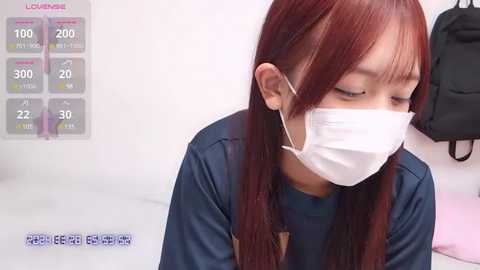 Media: Video of a young Asian woman with long red hair wearing a white surgical mask, blue shirt, and standing in a white room with a temperature gauge and thermometer display.