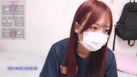 Media: A video of a young East Asian woman with long, straight red hair, wearing a white surgical mask and navy blue top, sitting in a sterile room with a black backpack hanging on the wall.