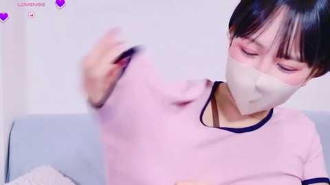 Media: Video of a young East Asian woman with short black hair, wearing a pink sweater and white face mask, sitting on a light blue couch. Background is plain white.