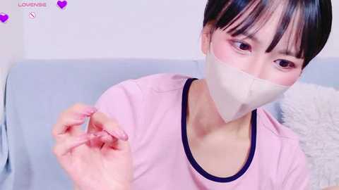 Media: Video of an East Asian woman with short black hair, wearing a pink t-shirt and white tape over her mouth, holding a small pink item.