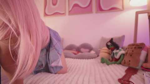 Media: Video of a young woman with long, straight, pink hair, kneeling on a white knitted blanket in a pastel-colored room. She wears a denim shirt and shorts, and a stuffed toy, resembling a green frog, is next to her.