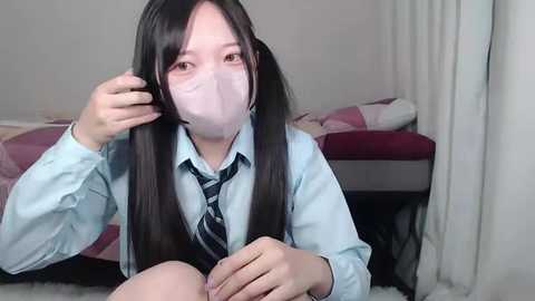 Media: Video of an East Asian woman with long black pigtails, wearing a light blue shirt, black tie, and white face mask, seated on a bed in a simple, dimly lit bedroom.