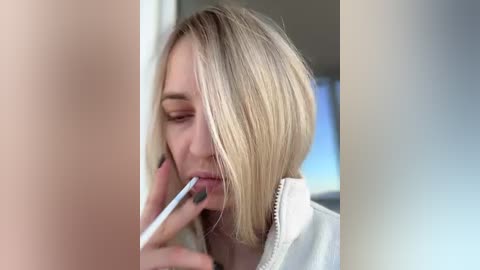 Media: Video of a blonde woman with a bob haircut, wearing a white jacket, applying lipstick with a silver tube, in a well-lit, modern bathroom setting.