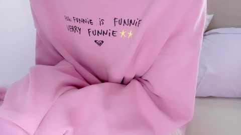 Media: Video of a person wearing a pink sweatshirt with \"I'm Funny\" and a heart symbol on it, lying on a bed with white sheets, in a cozy, intimate setting.