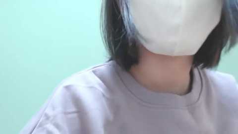 Media: Video of a person with short, straight black hair wearing a white surgical mask and a light grey t-shirt, against a plain light green background.