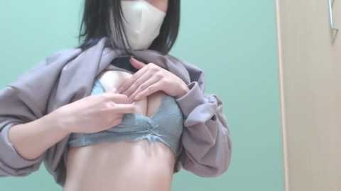 Media: Video of a slender, light-skinned woman with black hair, wearing a face mask, lifting a light gray hoodie to reveal a blue lace bra, exposing her small breasts.