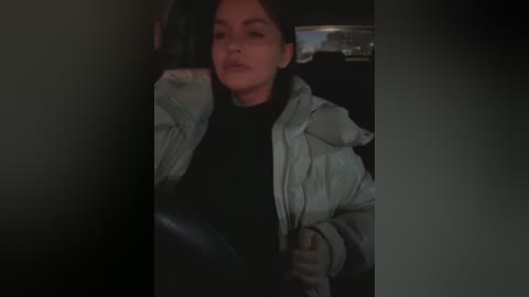 Media: Video of a woman with dark hair, wearing a black turtleneck and a light jacket, driving a car at night. She has a serious expression.