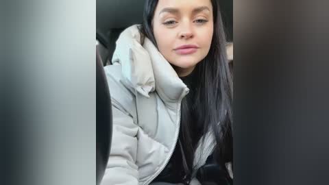 Media: Video of a young woman with straight black hair, wearing a puffy white jacket, sitting in a car. She has a serious expression. The background is blurry.