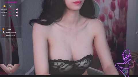 Video of a light-skinned woman with long, black hair, wearing a black, strapless lace top, seated indoors with a blurred floral backdrop.