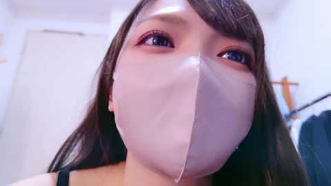 Media: Video of a young woman with long, straight brown hair, wearing a white surgical mask that covers her mouth and nose, in a well-lit, modern bathroom setting with white walls and a towel rack visible in the background.