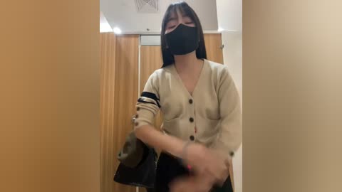 Media: A video shows an Asian woman with shoulder-length black hair and a black face mask, wearing a beige cardigan and holding a black bag. She stands in a hallway with beige walls and a wooden door.