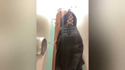 Media: Video of a woman in a tight, black leather skirt with lace-up back, standing in a public restroom. She has light skin and is bending forward, her buttocks prominent. The restroom has mint-green walls and a metallic toilet paper dispenser on the left.