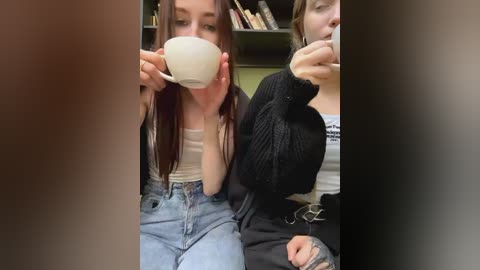 Media: Video of two young women, one in a beige tank top and blue jeans, the other in a black sweater, drinking coffee in a cozy indoor setting with bookshelves in the background.