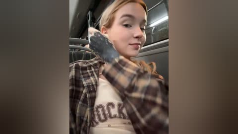 Media: A video of a young woman with fair skin and blonde hair, wearing a plaid shirt and a white T-shirt, sitting in a car with a grey interior. She's holding a black smartphone, and the background shows the car's window and ceiling.