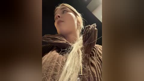 Media: Video of a blonde woman with fair skin, wearing a brown knitted cape, holding a strand of hair, looking upward in a dimly lit room.