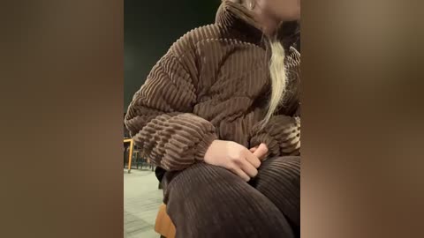 Media: Video of a person in a brown, textured sweater and pants, sitting with their head down, hands on their lap, in a dimly lit room with wooden furniture.