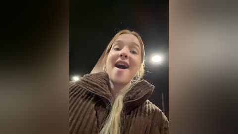 Media: Video of a young woman with long blonde hair, smiling broadly, wearing a brown, ribbed knit coat. The background is dark, with a single bright light source illuminating her face.