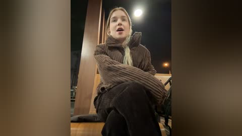 Media: A video of a young woman with blonde hair, wearing a brown knit sweater and black pants, sitting on a wooden bench, looking out a window at night.