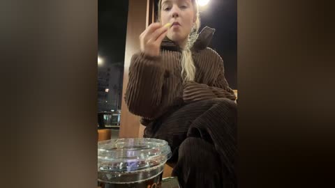 Media: A video of a blonde woman in a brown sweater, sitting indoors, holding a cigarette, with a glass of beer on the table in the foreground.