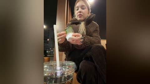 Media: Video of a blonde woman in a brown sweater, holding a white straw with a green lid, seated indoors, blurry background, wooden furniture, warm lighting.