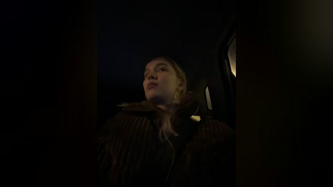 Media: A dimly lit video of a young woman with pale skin and blonde hair, wearing a brown jacket, looking upward. The background is dark and blurry, suggesting an intimate, nighttime setting.