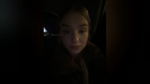 Media: A dimly lit video of a young girl with fair skin and light brown hair, wearing a brown coat, seated in a car.