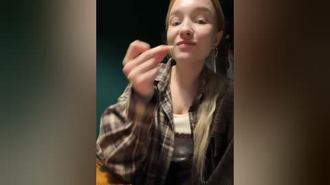 Media: Video of a young woman with fair skin and long blonde hair, wearing a plaid shirt and brown vest, holding a small object, against a blurred, dark background.