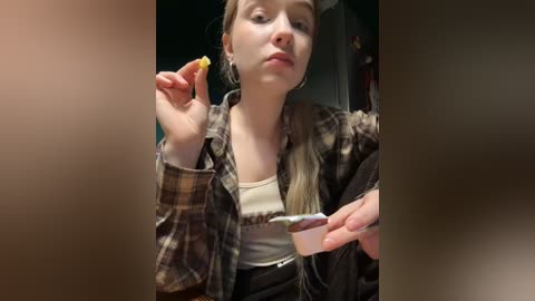 Media: Video of a young Caucasian woman with fair skin, wearing a brown plaid shirt over a white tank top, eating a cupcake and holding a yellow popsicle. Background is blurred.