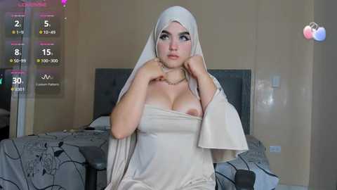 Media: Video of a young woman with light skin, wearing a white veil, revealing her large breasts, seated on a bed in a bedroom with a grey headboard and digital overlays.