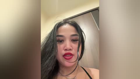 Media: Video of a young Asian woman with long black hair and red lipstick, captured from the waist up, wearing a black top, in a dimly lit room with beige walls and a wooden door.